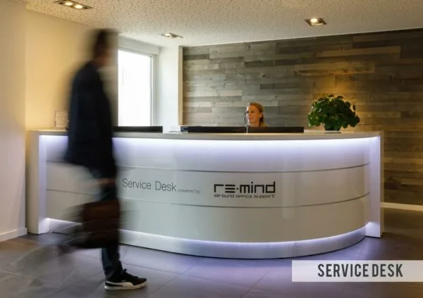 Receptiebalie van de Service Desk bij Re-mind aan de Baarnsche Dijk, met een medewerker achter de balie en een voorbijlopende persoon.
